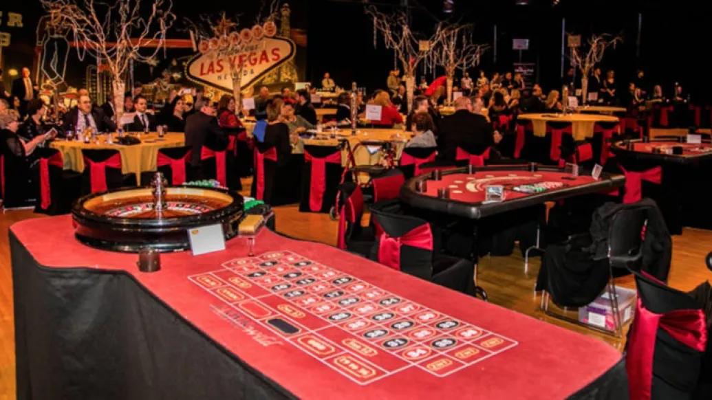 View of Casino Royale event with a roulette table in the foreground and additional casino tables surrounded by guests in the background.