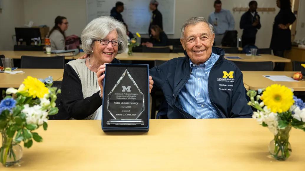 Susan and Arnold Coran are smiling and appear to have just received a plaque that Susan is holding toward the viewer. They are seated behind a table decorated with two vases of flowers.
