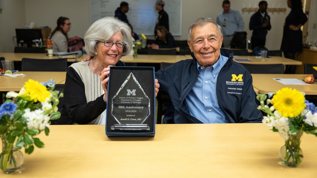 Susan and Arnold Coran are smiling and appear to have just received a plaque that Susan is holding toward the viewer. They are seated behind a table decorated with two vases of flowers.