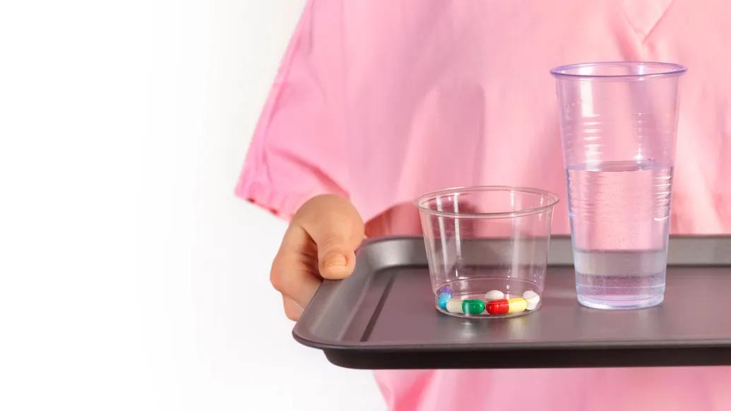 pills in a cup water in a cup both on a tray with a patient in pink gown
