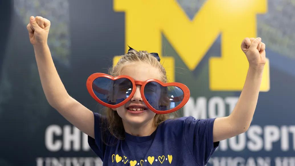 Paisley, a young girl with long brown hair that is pulled back on top, is wearing oversized heart-shaped red sunglasses and a blue Save A Heart t-shirt with yellow hearts across the top. She is standing with her arms extended upwards in a gesture of victory in front of a block M.