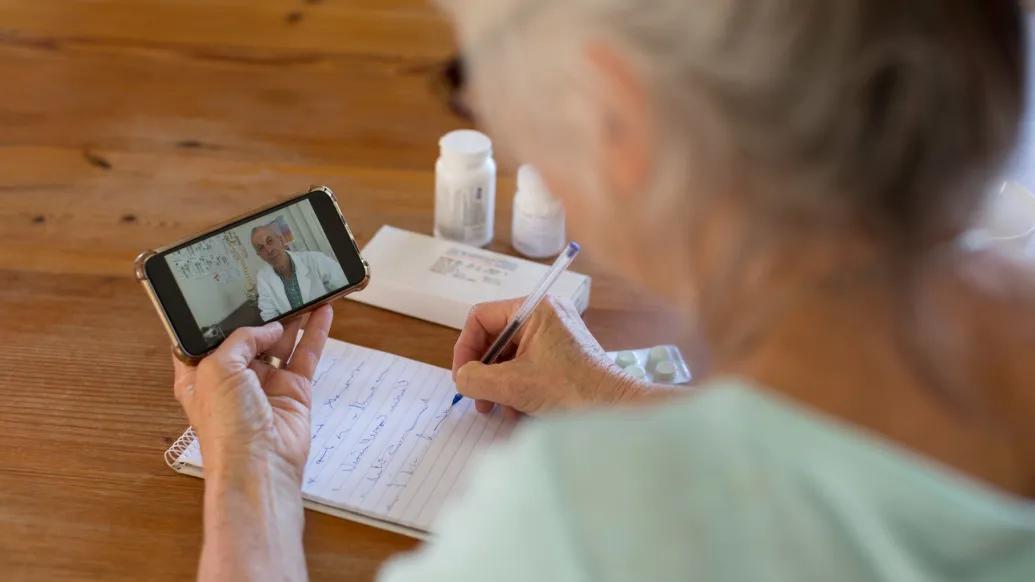 An older woman seen from behind holds a phone on which a doctor is talking to her while she takes notes with pill bottles nearby 