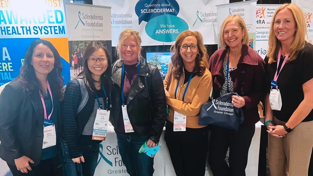 six women standing together smiling in front of posters