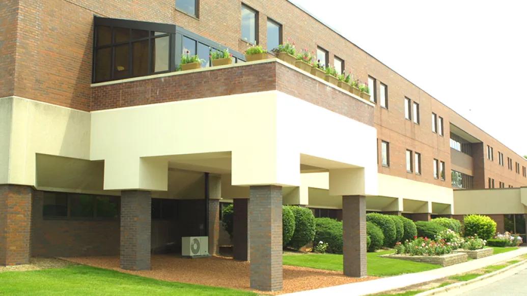 Ground level view of the entrance of Sparrow Hospice House. It is a 3-story brick and concrete building with an extension over the entrance. There are leavy bushes and flowers in front of the building.