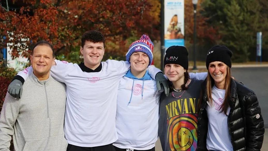 Marc, Brody, Max, Caroline, and Alyssa Tushman standing with their arms over each others' shoulders and smiling toward the camera. They're wearing cold weather gear and fall foliage is visible behind them.