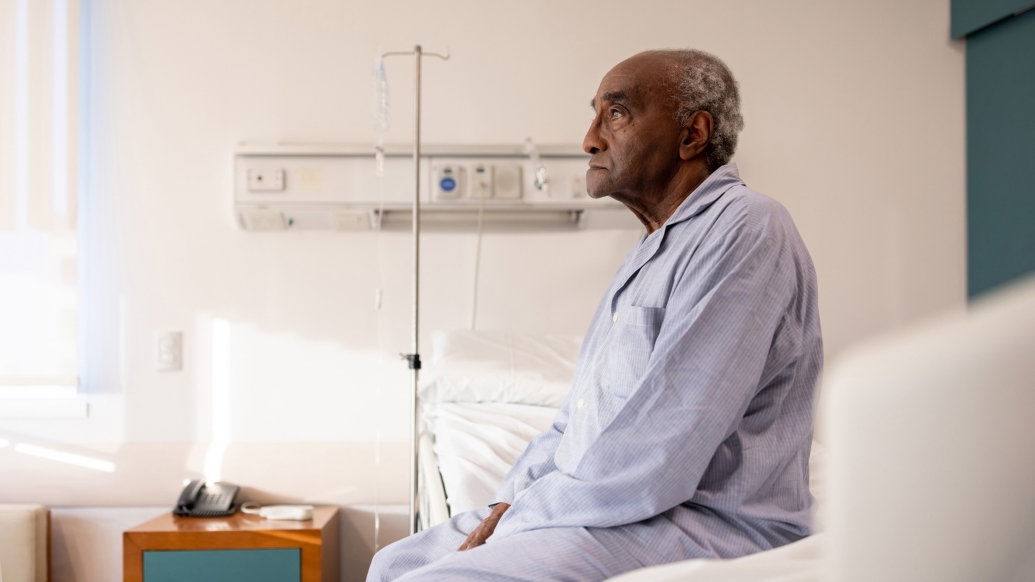 man on white bed in blue pajamas with iv pole behind him neutral expression on face