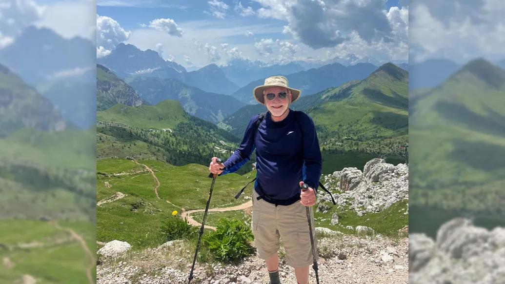 man hiking navy long sleeve shirt and short tan pants mountains sky behind him