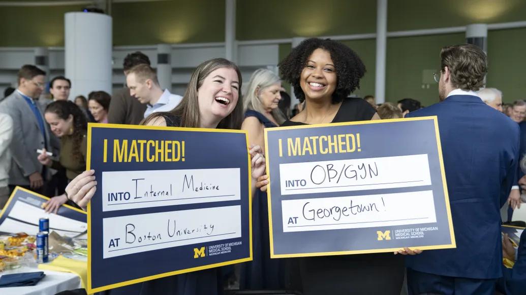 Two U-M medical school students hold up cards announcing their residency matches. The left card reads "Internal Medicine at Boston University." The right card reads "OB/GYN at Georgetown!"