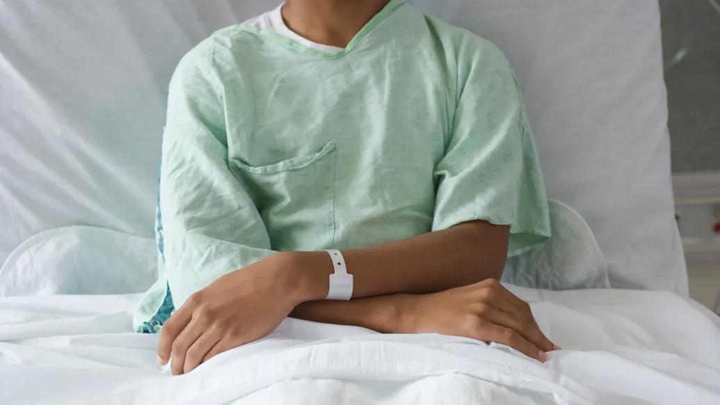 The torso and arms of a thin brown-skinned adolescent male in a hospital gown and bed