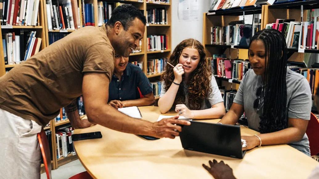 teacher with students at table looking at laptop in library 