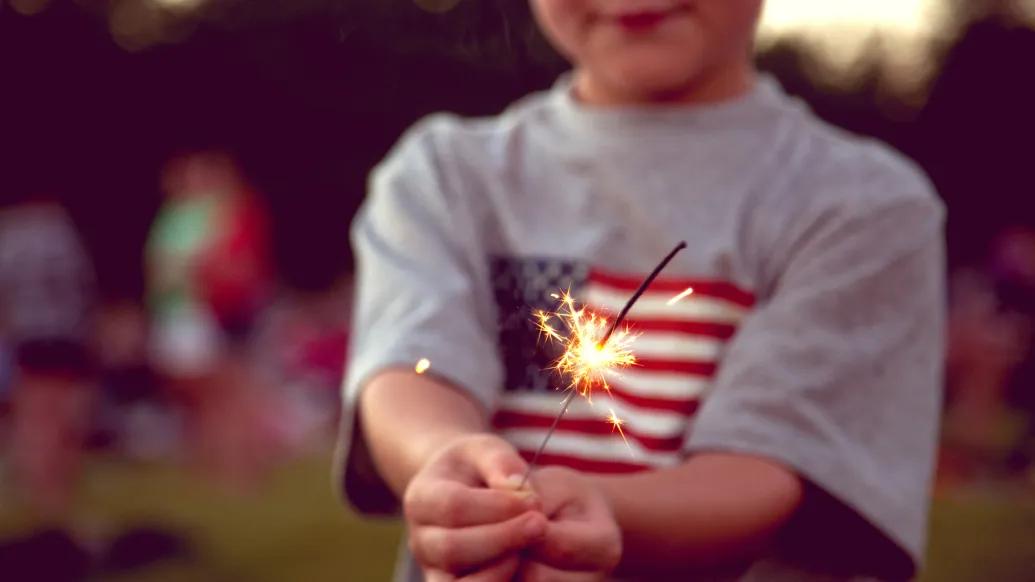 Little boy in American flag t-shirt holds a sparkler near the end of it’s time burning in his hands 
