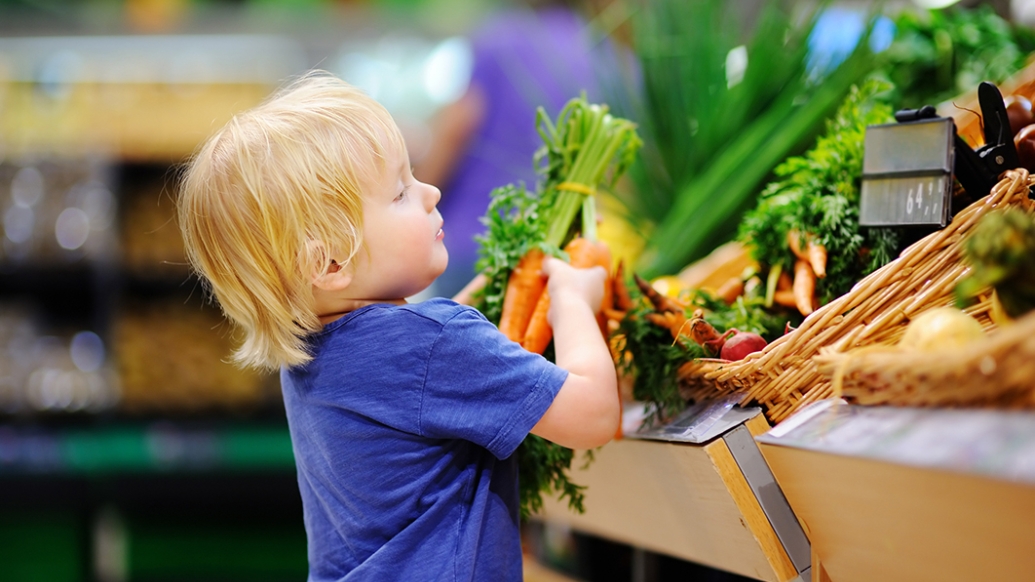 A small child in a blue t-shirt picks up a bunch of carrots from a produce display.