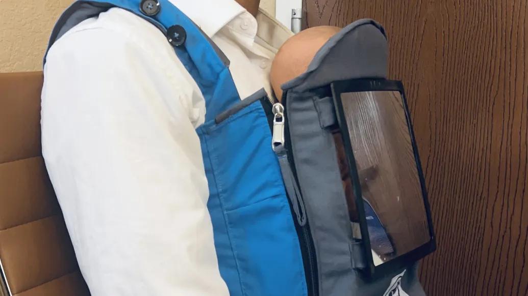 A person wears a protype infant sling on the front of their body. The sling has a special window to allow light to reach the baby.
