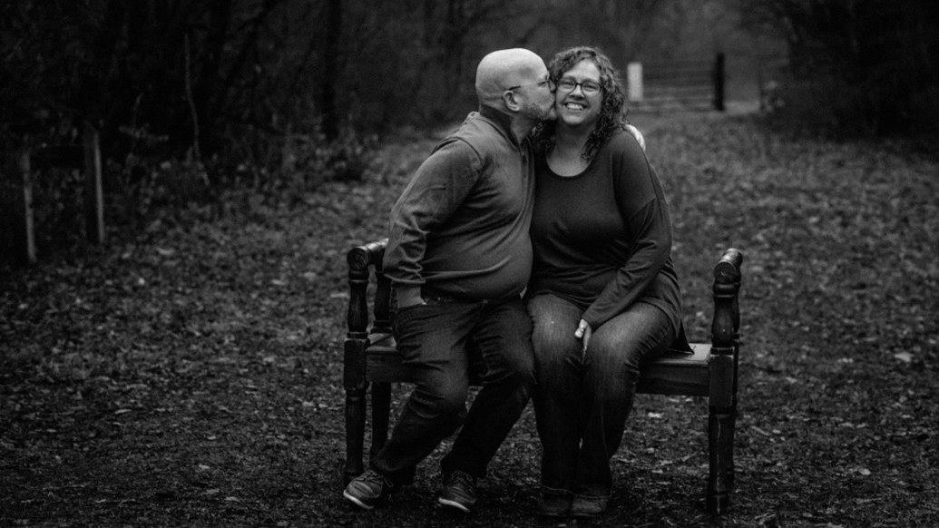 couple sitting on bench black and white kissing on cheek 