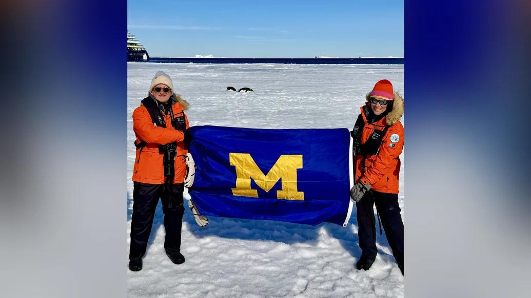 Bob and Debbie Merion, wearing orange parkas, stand on the Antarctic ice holding a U-M flag between them.