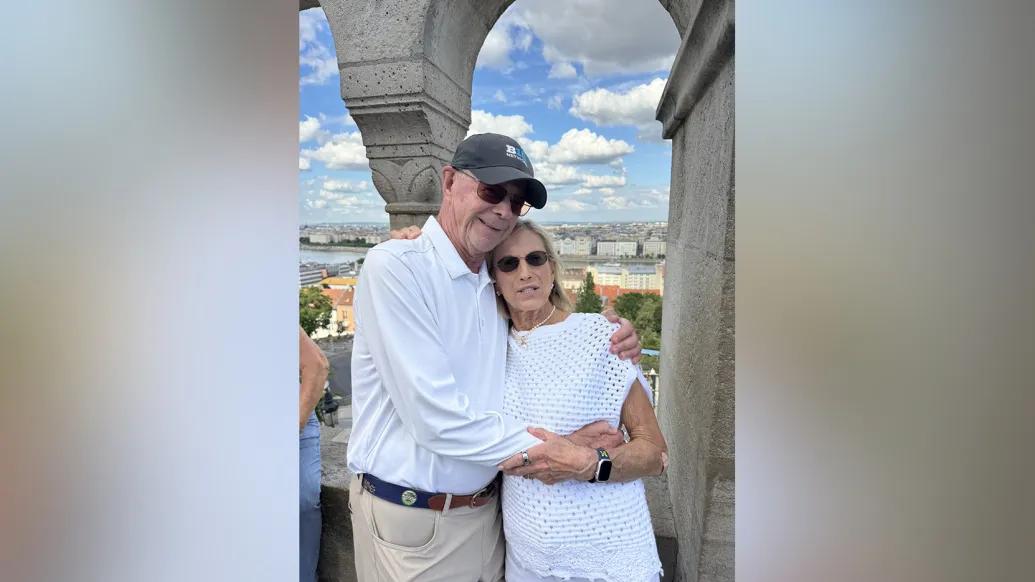 Bill and Sally Martin stand affectionately with their arms around each other. They appear to be in a high open tower above a city.