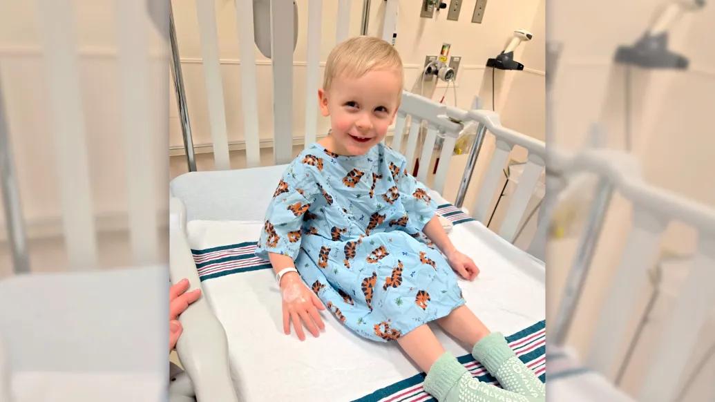 Young boy in hospital bed smiling, wearing blue hospital gown with tigers all over it and green hospital socks