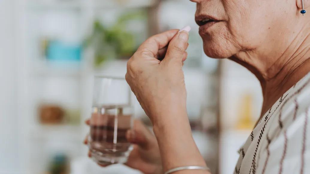 A person holds a pill near their mouth and a glass of water in the other hand, preparing to take the medication. The background is a softly focused room with shelves and blurred greenery.