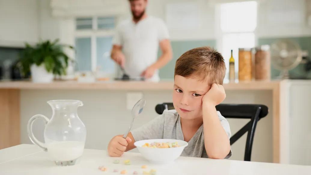 kid at table not wanting to eat breakfast with parent blurred in background white coloring milk on table