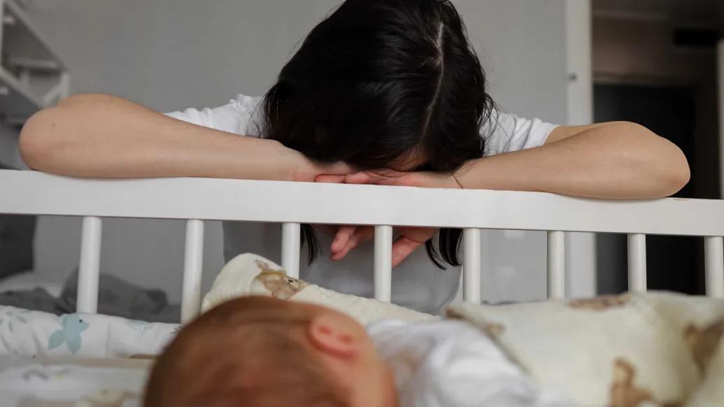 A dark haired woman with her head resting on her arms face down on the rail of a crib with a baby visible in the foreground