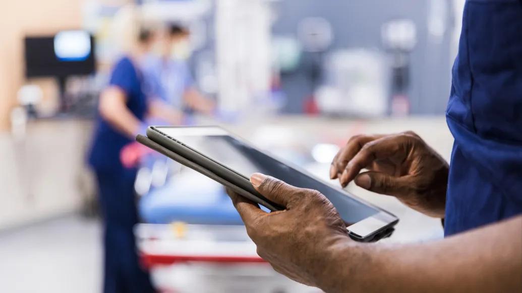 Two people in medical scrubs are in a blurred hospital setting. In the foreground, hands are holding a digital tablet.