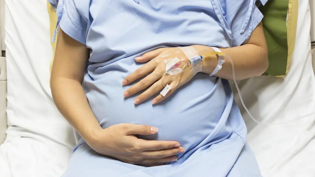 Woman's torso in a blue hospital gown, with her hands holding a pregnant belly and an IV in place.