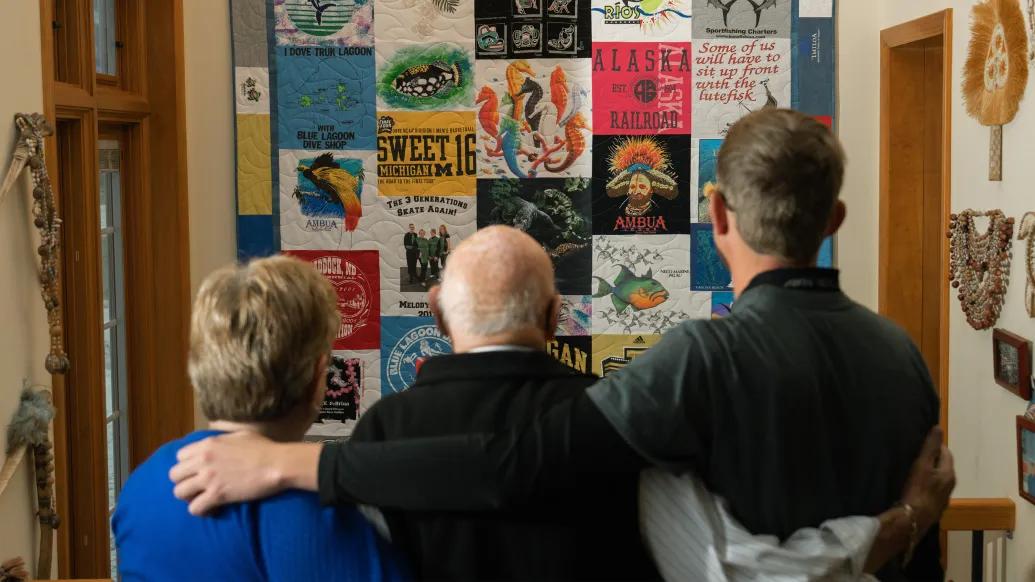 Three people stand arm in arm with backs to camera studying a wall of family mementos