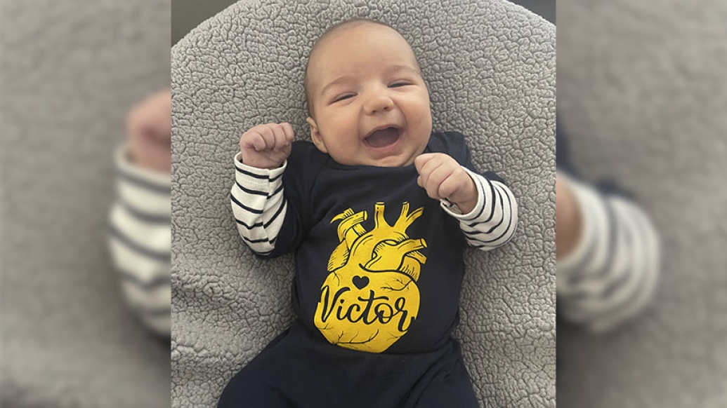 baby smiling on grey pillow with blue and yellow shirt on and striped undersleeve shirt