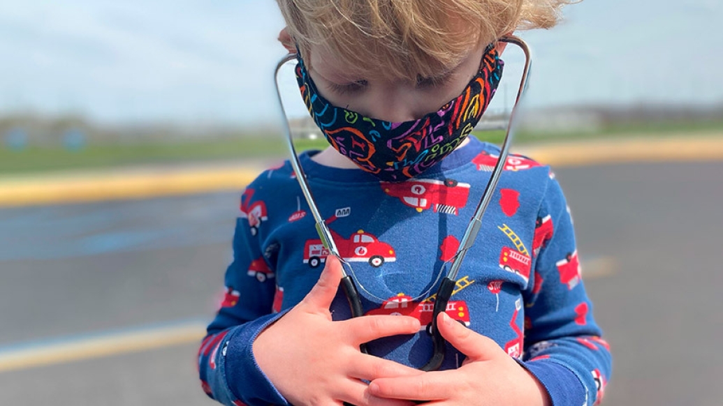 child looking down at stethoscope on his chest while wearing mask outside and wearing blue shirt with firetrucks on it