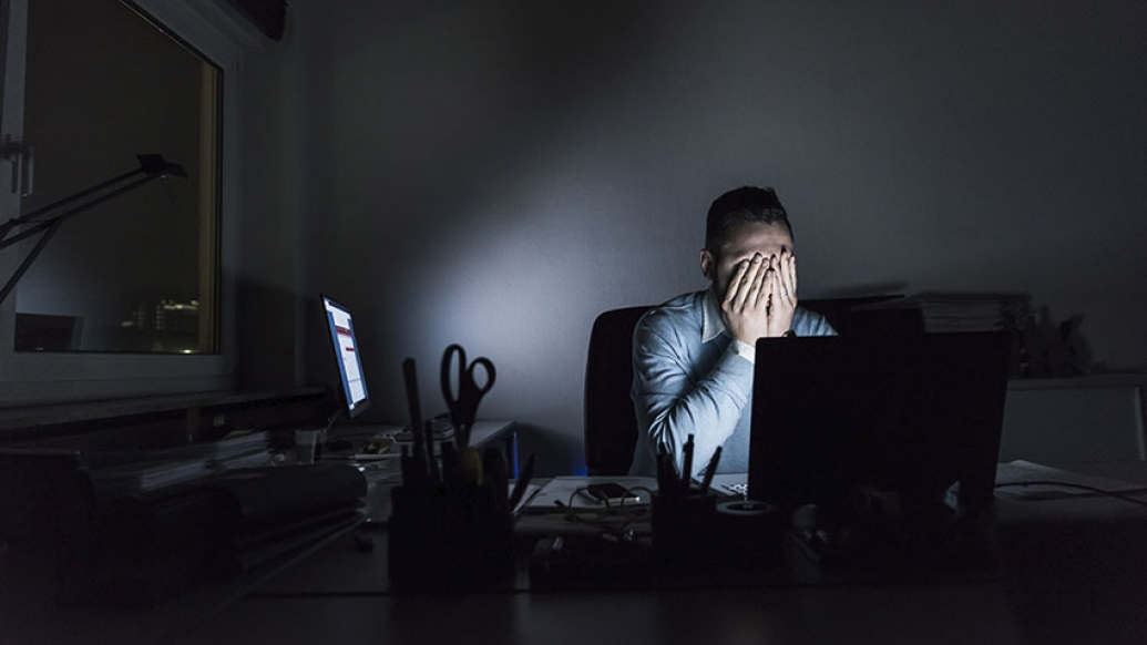 Man sitting behind a computer in a darkened room with his hands over his face