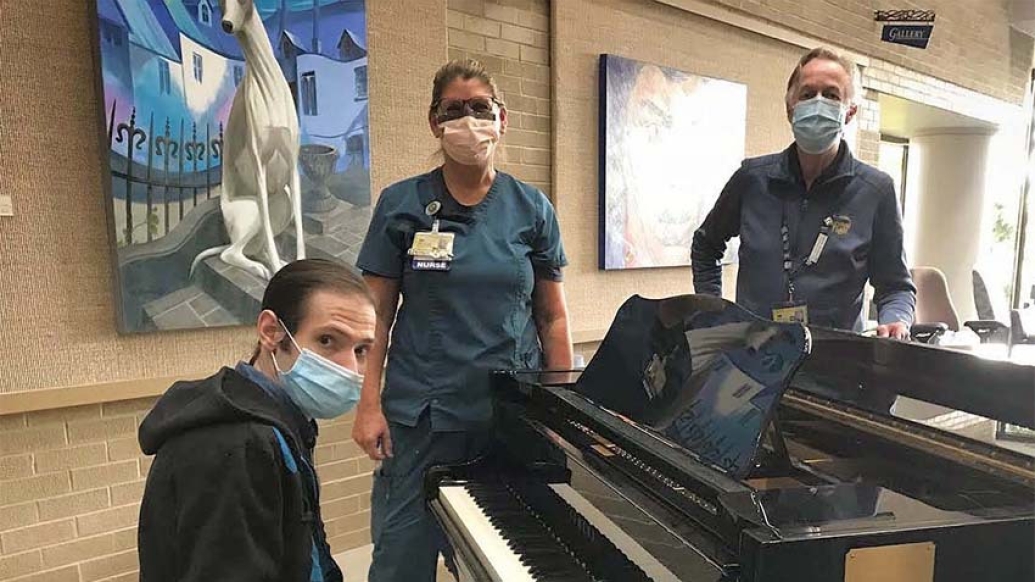 man sitting at piano with mask on and nurse with mask on standing near him and man with mask on standing across piano