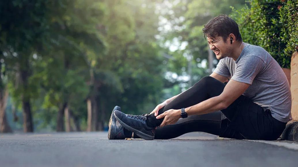 Man sitting on sidewalk among green trees holding his hurt ankle