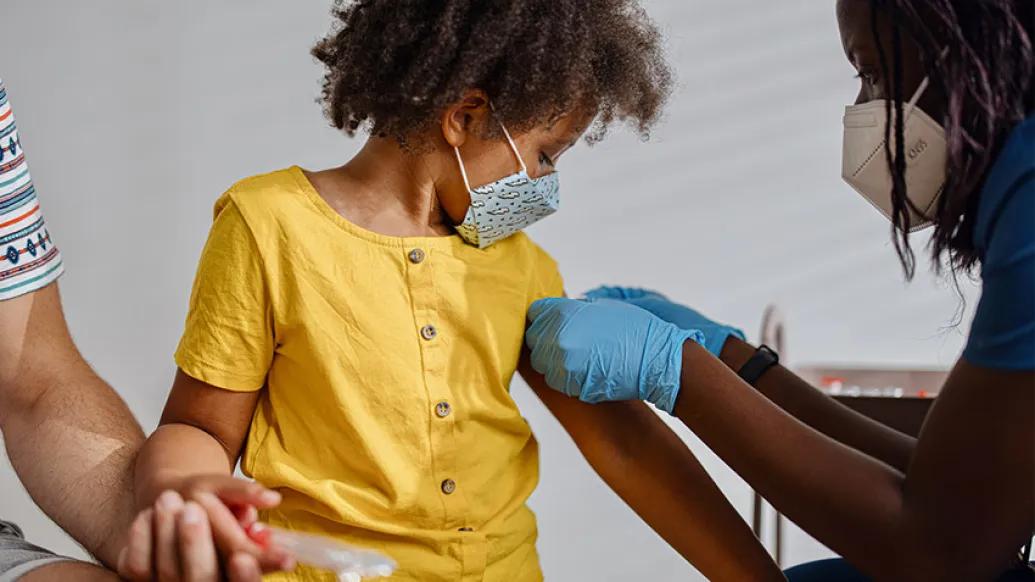 child in yellow shirt sitting looking down on arm with blue mask on and nurse with globes putting band-aid on