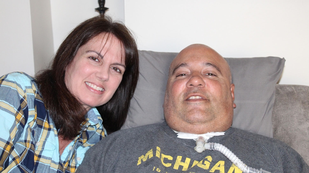 woman with dark hair standing next to man with tracheostomy sitting in chair