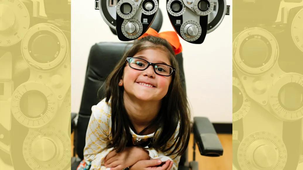A young girl with glasses smiles while sitting in an eye exam chair under a phoropter.