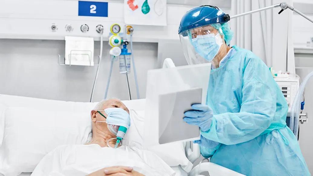 older patient in white hospital gown in ICU with doctor in blue protective gown and equipment and face shield holding ipad up
