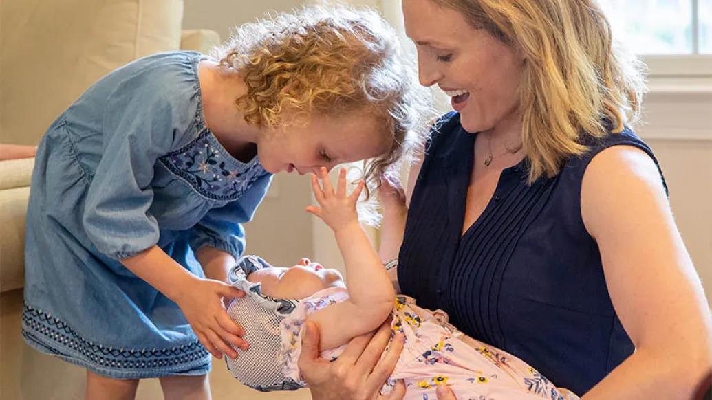 Mother in blue sleeveless top holding and look down at her baby with blue hat and pink outfit with flowers and toddler also looking at baby wearing blue dress