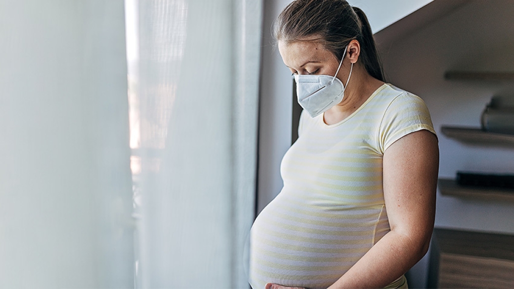 pregnant mother wearing a mask indoors looking at her belly 