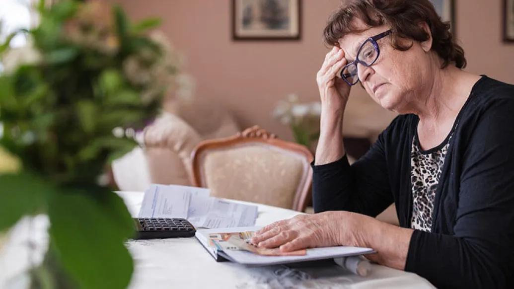 Senior woman with hand on head looking at book with calculator next to her at table