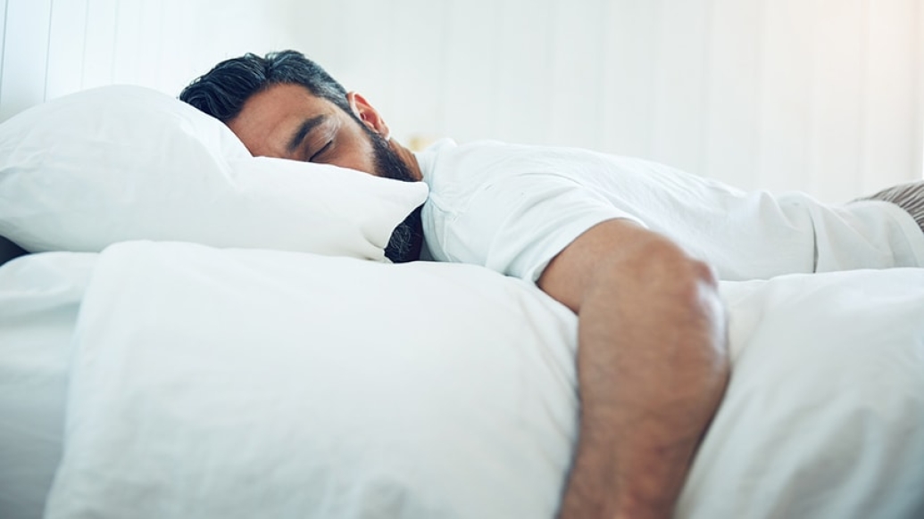 A man with a beard lies peacefully asleep on a bed, surrounded by soft white bedding.