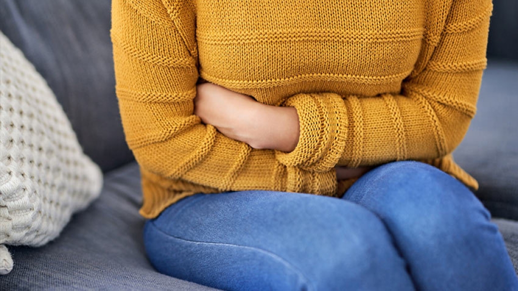 woman holding stomach pain mustard colored shirt jeans sitting on couch