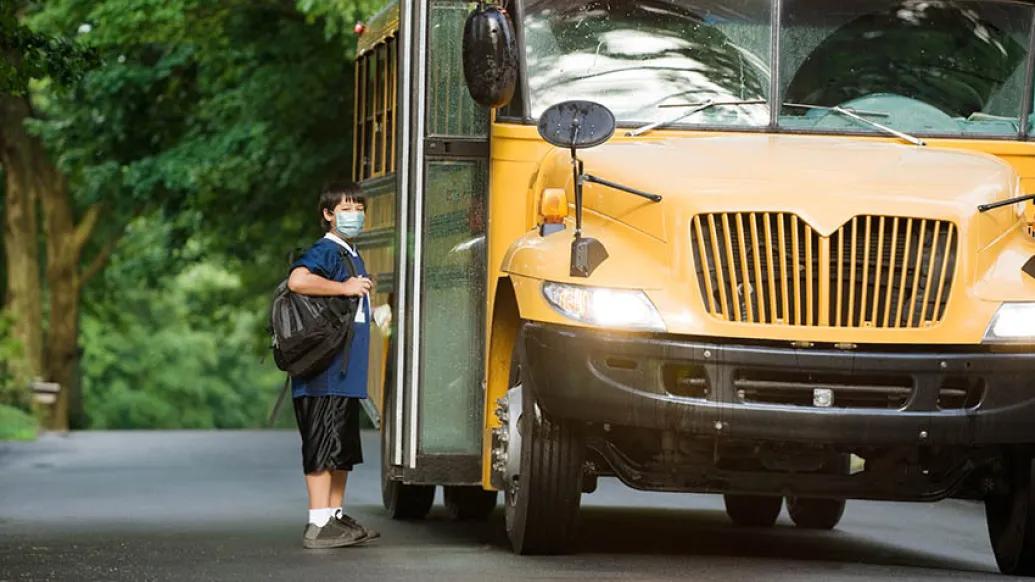 Child with brown hair and mask on in front of open doors of school bus wearing backpack