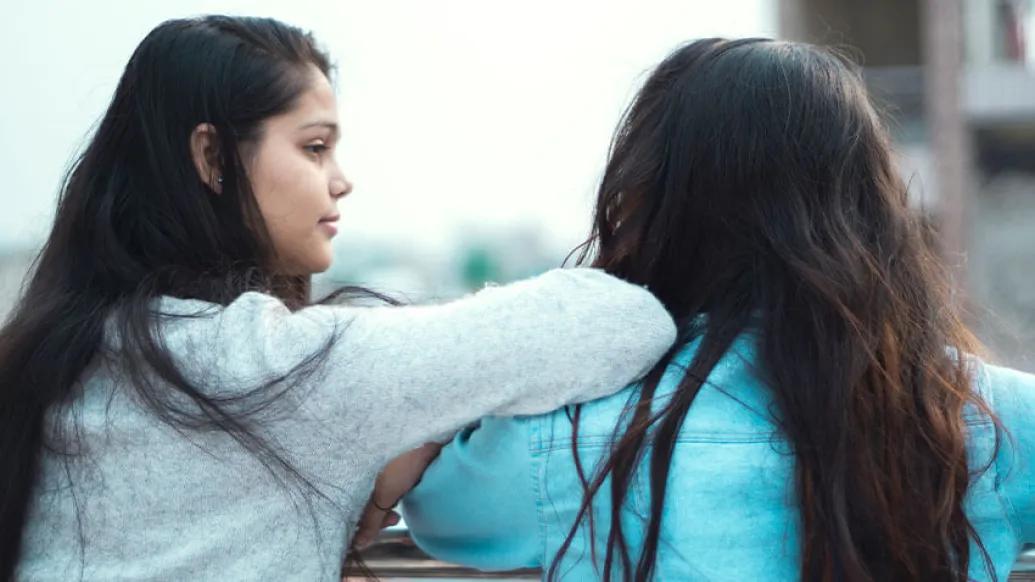 girl with long black hair looking and leaning on friend's shoulder with long brown hair and blue shirt