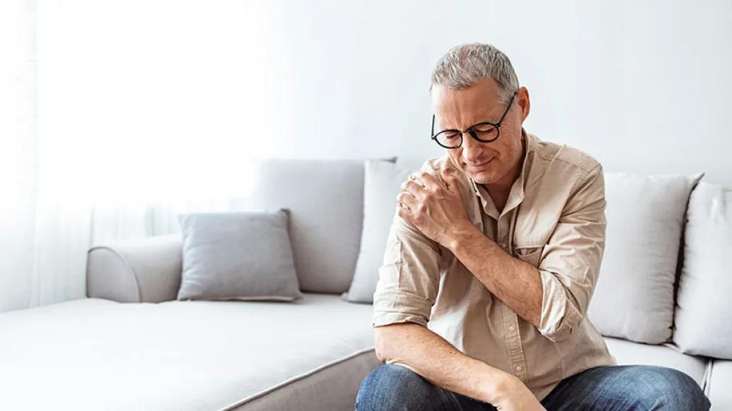 Man with glasses sitting on edge of furniture leaning, holding his shoulder.