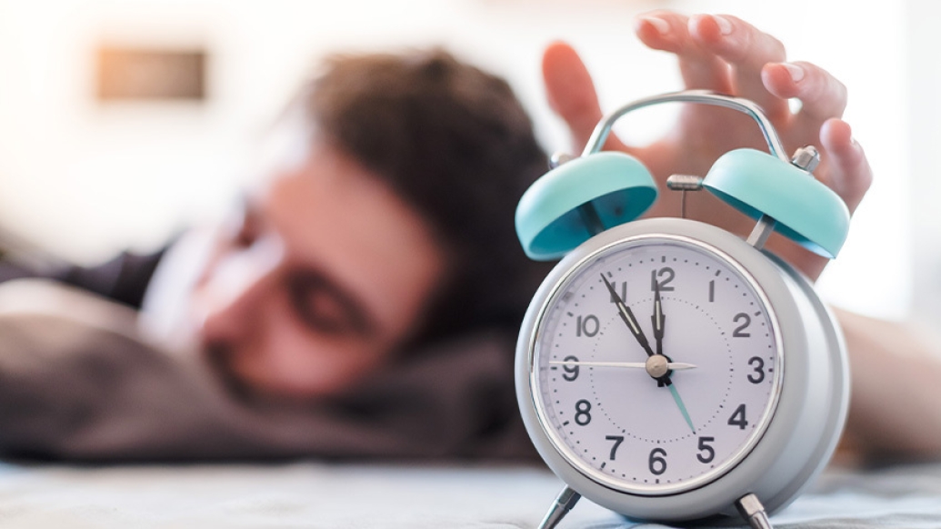 man sleeping reaching to shut off blue and white alarm clock