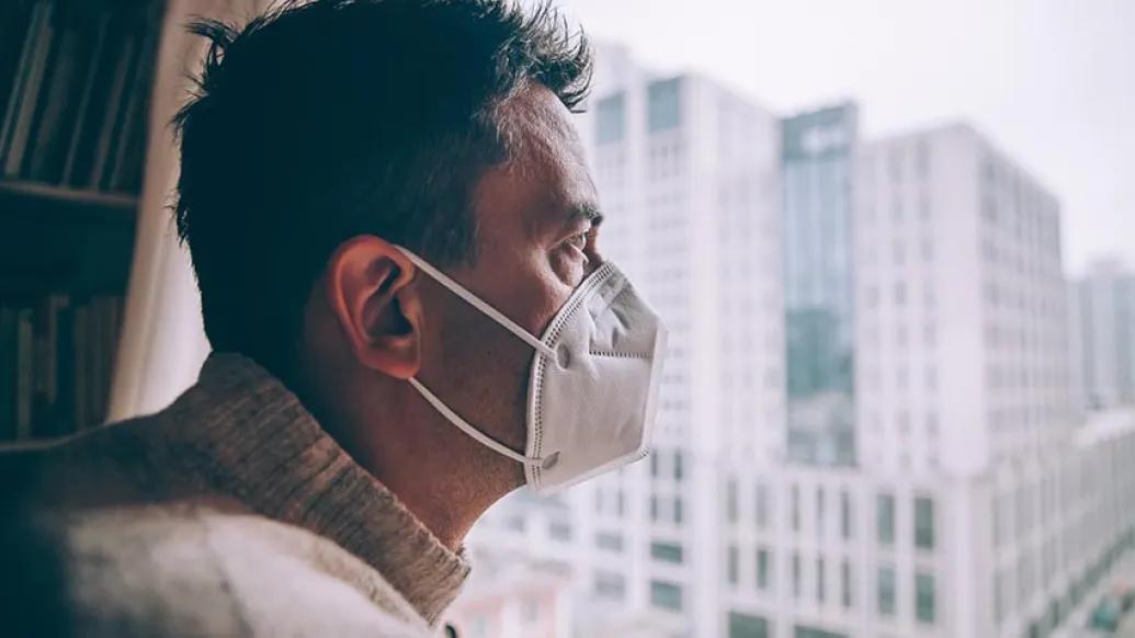 Man with mask on staring outside the window looking at city.