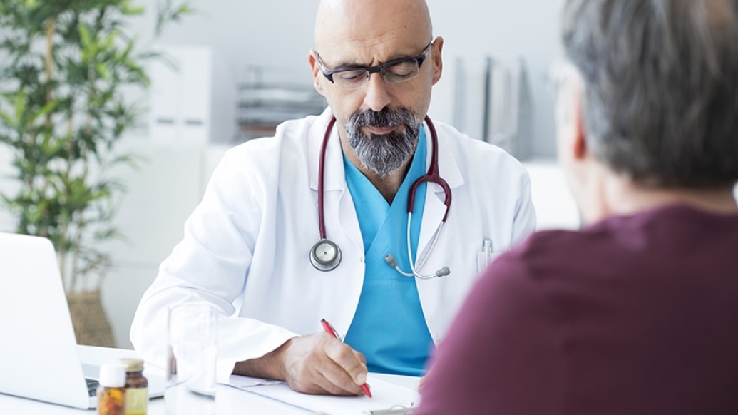 doctor with white coat sitting with stethoscope around his neck and blue shirt talking with patient with short grey hair and maroon shirt