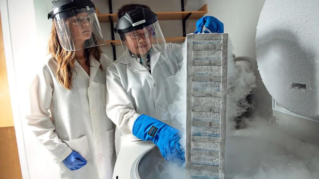 two scientists in a lab with white coats and face shields with blue gloves 