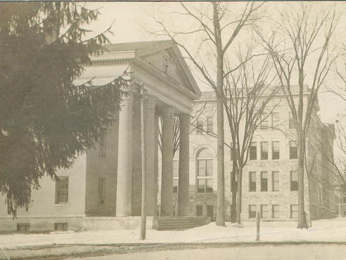Old photo of building with columns with bare trees, snow on the ground, and an evergreen tree in the foreground
