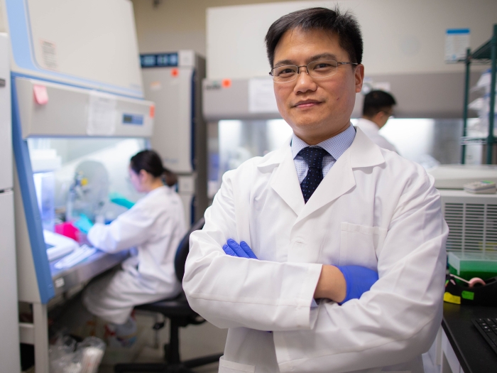 Asian main in white lab coat and blue surgical gloves standing with arms folded with lab n background